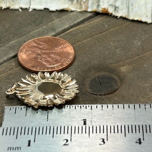 May include: A silver-colored pendant with a floral design, next to a penny and a ruler. The pendant is approximately 0.75 inches in diameter. The ruler shows measurements in millimeters.