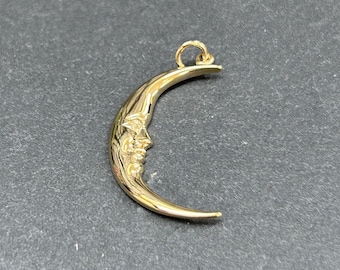 May include: A gold-colored crescent moon pendant with a detailed face design. The pendant has a loop at the top for a chain and is set against a dark gray background. The moon's face is intricately carved.