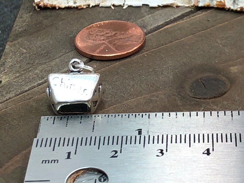 May include: Silver charm in the shape of a Chinese takeout container. The charm is about 1 inch tall and is sitting on a ruler. The charm has the word "Chinese" engraved on the side.