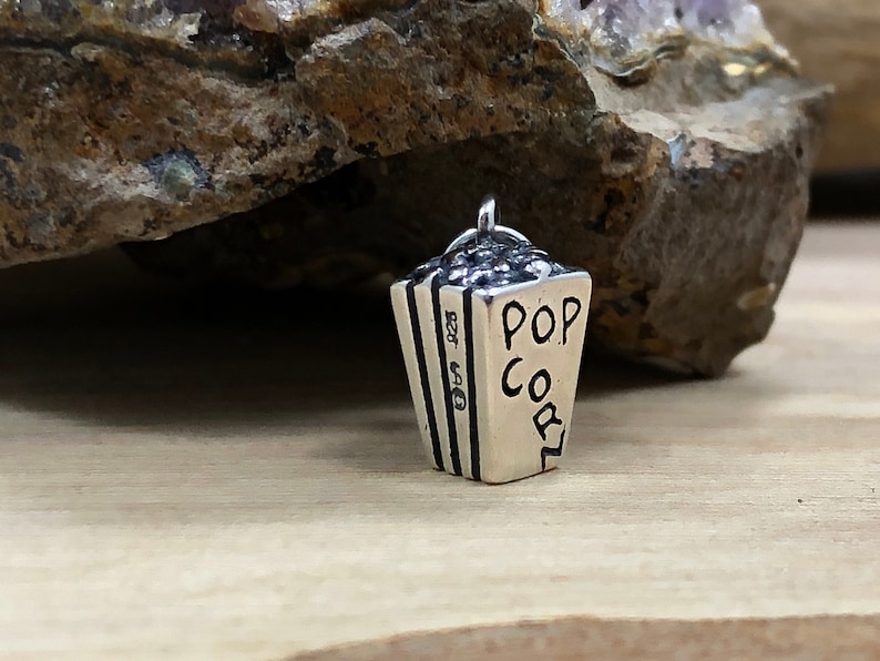 Może przedstawiać: Srebrny charm w kształcie wiaderka na popcorn z napisem "POP CORN" na boku.