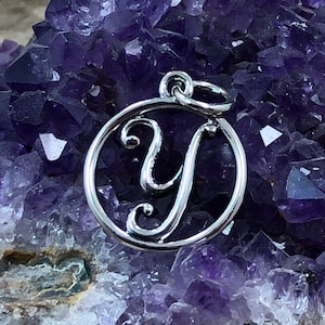 May include: Silver charm with a cursive letter Y in a circle, on a background of purple amethyst crystals.