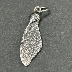 May include: A silver-colored pendant in the shape of a maple seed, also known as a helicopter seed. The pendant has detailed veining and a small loop at the top for a chain. The background is a dark gray.