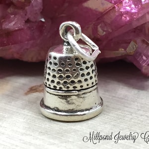 May include: A silver thimble charm with a textured surface. The charm is designed to look like a traditional thimble and has a loop for attaching to a necklace or bracelet.
