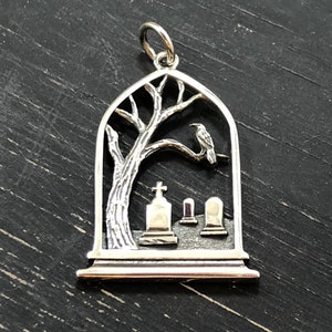 May include: Silver pendant charm featuring a detailed silhouette of a tree with a crow perched on a branch, set against a backdrop of three small tombstones. The pendant is shaped like an arched window.
