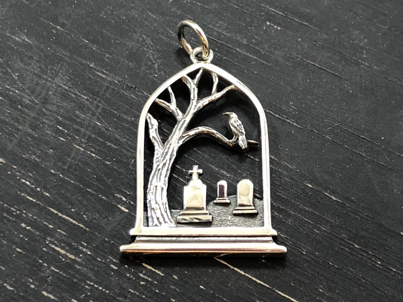 May include: Silver pendant charm featuring a detailed silhouette of a tree with a crow perched on a branch, set against a backdrop of three small tombstones. The pendant is shaped like an arched window.