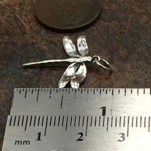 May include: A small, silver dragonfly charm with detailed wings and body. The charm is approximately 1 inch long, as measured by the ruler in the image. The dragonfly is set against a dark, textured background.