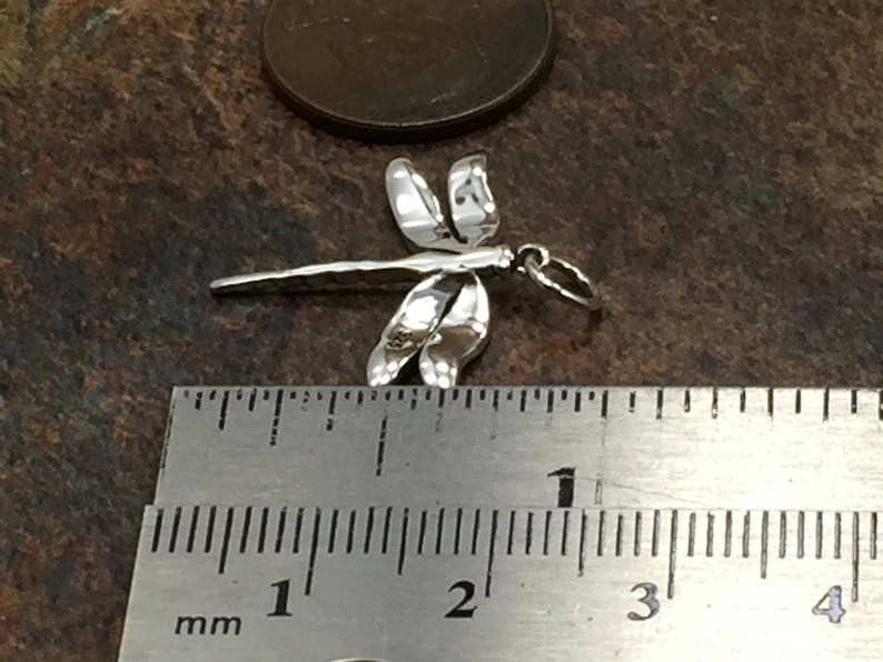 May include: A small, silver dragonfly charm with detailed wings and body. The charm is approximately 1 inch long, as measured by the ruler in the image. The dragonfly is set against a dark, textured background.