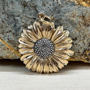 May include: A gold-colored sunflower pendant with a detailed design. The pendant features a textured center and layered petals. The pendant is designed to be worn as a necklace.