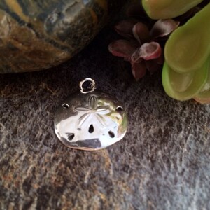 May include: A polished silver-toned sand dollar pendant featuring a five-pointed star pattern embossed on the surface, with small circular cutouts and a small metal jump ring attached to the top for hanging as a piece of jewelry.