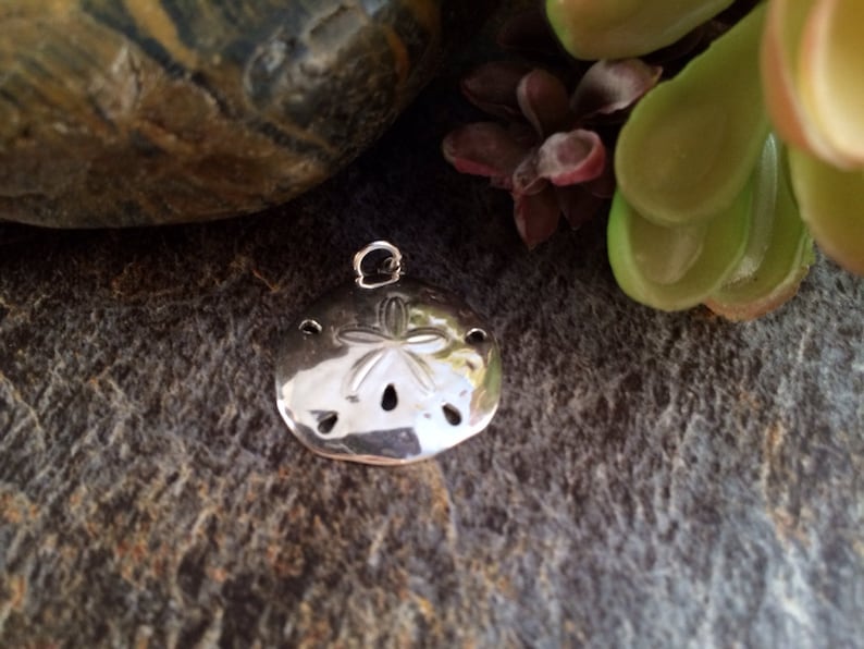 May include: A polished silver-toned sand dollar pendant featuring a five-pointed star pattern embossed on the surface, with small circular cutouts and a small metal jump ring attached to the top for hanging as a piece of jewelry.