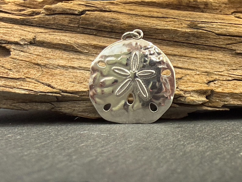 May include: A polished silver-toned pendant shaped like a sand dollar, featuring a central five-pointed star pattern and five small circular perforations. The pendant includes a small circular jump ring at the top for attachment to a necklace chain.