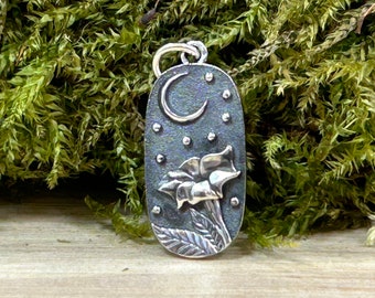 May include: A silver pendant with an oval shape, featuring a crescent moon and stars at the top. Below, a detailed flower design is visible. The pendant has a textured, dark background and a silver bail for a chain.