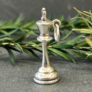 Puede incluir: Un amuleto de rey de ajedrez de plata con un bucle para sujetarlo a un collar o pulsera. El amuleto es detallado y tiene un acabado texturizado.