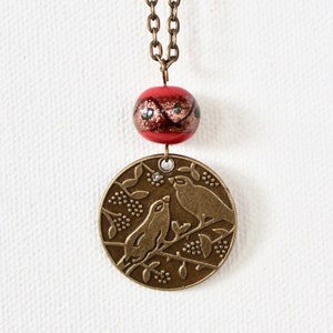 May include: A bronze necklace with a round pendant featuring two birds perched on a branch with leaves. The pendant is attached to a red and gold bead.