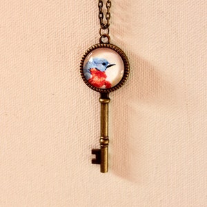 May include: A gold key necklace with a round glass pendant featuring a bluebird with a red breast. The pendant is attached to a gold key charm.