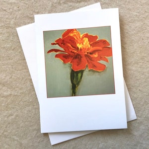 May include: A greeting card featuring a vibrant orange marigold flower painting. The flower has a yellow center and green stem, set against a muted gray background. The card is bordered in red and comes with a white envelope.