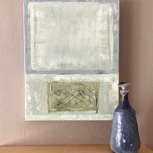 May include: Abstract painting with a textured green and white grid pattern in the lower portion of the canvas. The painting is framed in a white frame and is hanging on a light pink wall. A blue ceramic bottle with a white stopper is sitting on a light brown wooden surface.