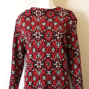 May include: A long-sleeved sweater with a striking geometric pattern in red, black, and white. The design features a repeating motif of diamond shapes and intricate details, creating a visually appealing and bold look. The sweater is displayed on a mannequin.