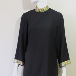 May include: A black mini dress with gold and green trim around the collar and cuffs. The dress has a mock neck and long sleeves.