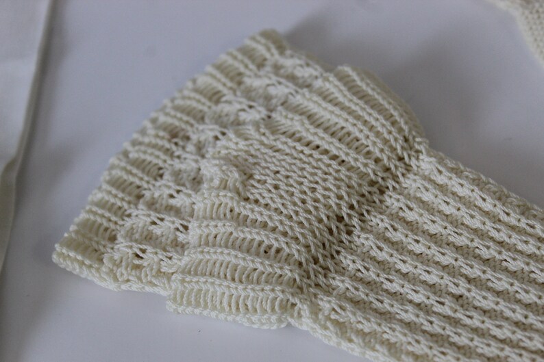 May include: Close-up of a cream-colored knitted fabric with a textured pattern. The fabric is soft and has a delicate, open weave.