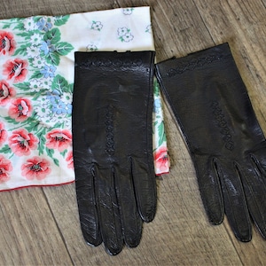 Könnte beinhalten: Ein Paar schwarze Lederhandschuhe mit dekorativen Nähten. Die Handschuhe liegen auf einem weißen Stoff mit Blumenmuster.