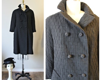 May include: A black textured coat with a double-breasted design and a notched collar. The coat has three-quarter sleeves and decorative buttons. A mannequin head with a hat is visible in the background.