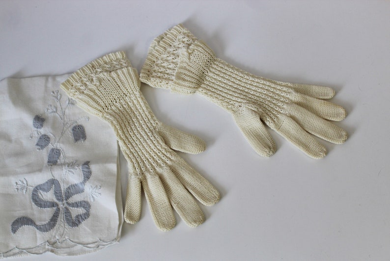 May include: A pair of off-white knitted gloves with a delicate lace pattern. The gloves are long and extend past the wrist.