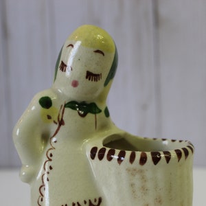 May include: A ceramic figurine of a woman with yellow hair, green eyes, and a brown dress. She is holding a small, white planter with brown trim.