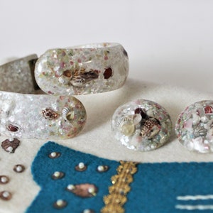 May include: A white plastic bangle bracelet and a pair of earrings with embedded seashells and iridescent glitter.