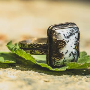 Può includere: Un anello in stile vintage con una pietra rettangolare bianca e nera incastonata in metallo color bronzo. L'anello poggia su una foglia verde, mostrando intricati motivi a foglie e geometrici. Il design evoca un'estetica bohémien o ispirata alla natura.