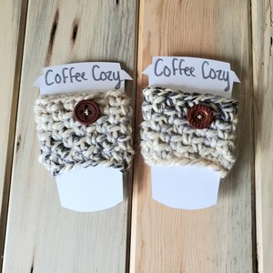 May include: Two white crocheted coffee cup cozies with brown buttons. The cozies are on a wooden background with a white card that says "Coffee Cozy" on each.
