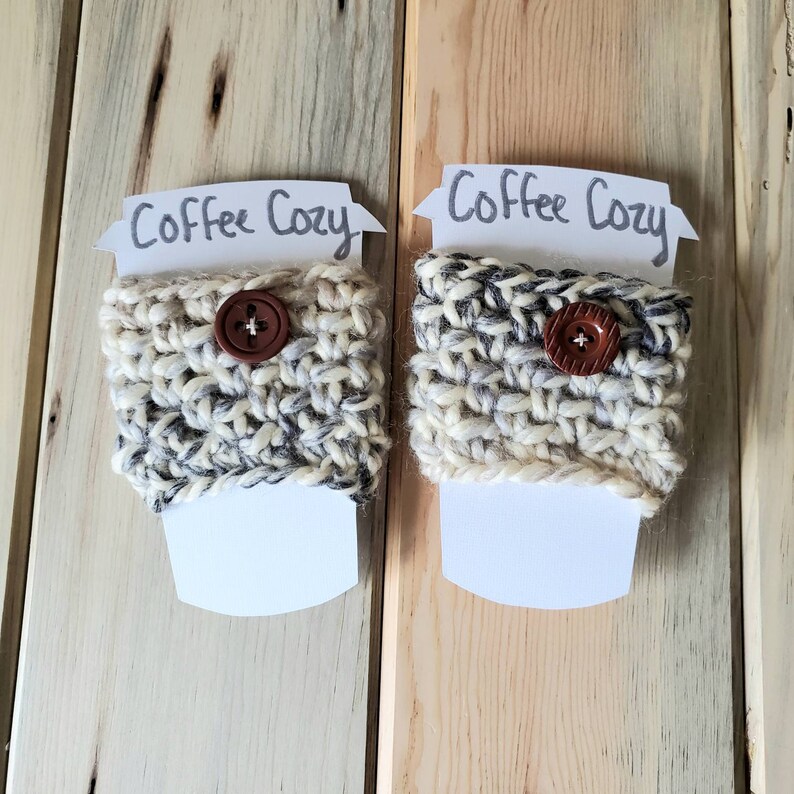 May include: Two white crocheted coffee cup cozies with brown buttons. The cozies are on a wooden background with a white card that says "Coffee Cozy" on each.