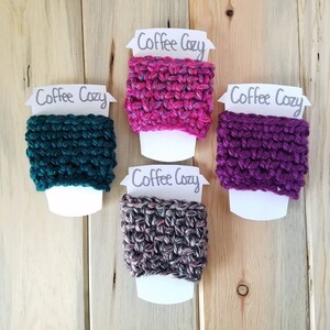 May include: Four crocheted coffee cup cozies in different colors: teal, pink, purple, and gray. Each cozy is displayed on a white card with the text "Coffee Cozy" written in gray.