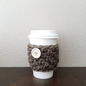 May include: A brown and white crocheted coffee cup cozy with a white button. The cozy is on a white paper cup.