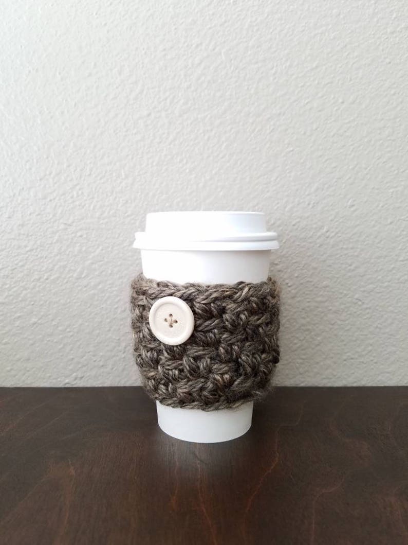 May include: A brown and white crocheted coffee cup cozy with a white button. The cozy is on a white paper cup.