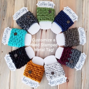 May include: A variety of crocheted coffee cup cozies in different colors, including teal, green, gray, black, brown, burgundy, gold, and white. Each cozy has a metal tag that says "Coffee Cozy". The cozies are arranged in a circle on a wooden surface. Customize a Hand Stamped Metal Tag!