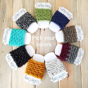 May include: A selection of ten crocheted coffee cup cozies in various colors, including teal, gray, green, navy, brown, burgundy, black, white, and yellow. The cozies are arranged in a circle on a wooden surface. The text "Pick your colors" is written in the center of the circle.