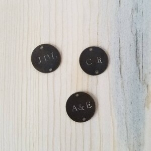 May include: Three round metal tags with initials engraved on them. The tags are dark brown and have two holes on each side. The initials on the tags are JDL, CR, and A&E.