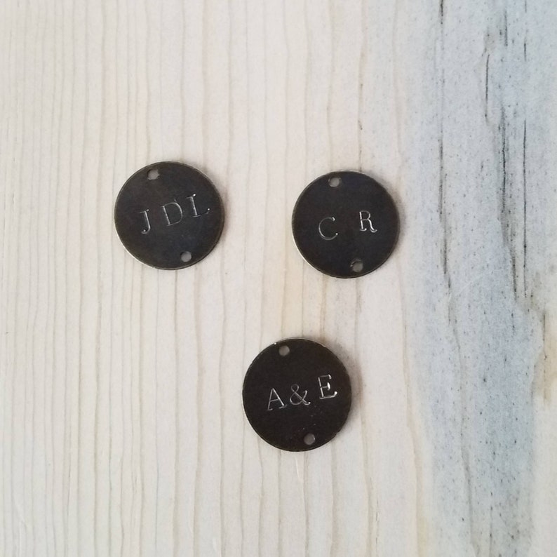 May include: Three round metal tags with initials engraved on them. The tags are dark brown and have two holes on each side. The initials on the tags are JDL, CR, and A&E.