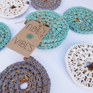 Op de afbeelding: Set van zes gehaakte onderzetters in grijs, wit en turquoise. De onderzetters zijn gemaakt van touw van natuurlijke vezels en hebben een ronde vorm. Een bruin label met de tekst "Fiber Vibes" is bevestigd aan de onderzetters.