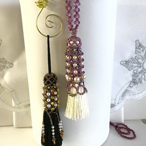 May include: Two beaded tassels with white and black fringe hanging from a white stand. The tassels are decorated with purple, gold, and white beads.