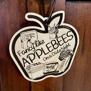 May include: A wooden apple-shaped ornament with the text "Fancy like Applebees on a Date Night". The ornament features a black and white illustration of a cooler, a drink, a box of snacks, and a box of matches.