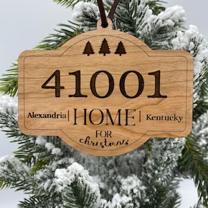 May include: Wooden Christmas ornament with a house-shaped design. It features the text "41001 HOME" with "Alexandria" and "Kentucky" on either side. The ornament also includes the text "FOR christmas" and three small pine trees.