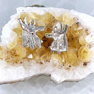 May include: Two silver Anubis charms, one facing left and one facing right, are displayed on a bed of yellow citrine crystals.