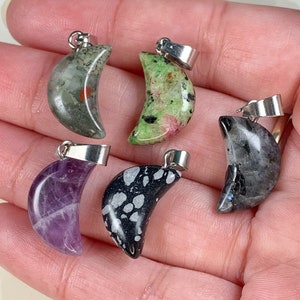 May include: Six crescent moon shaped pendants made of various stones. The stones are green, black, purple, and grey with white speckles. The pendants are attached to silver jump rings.