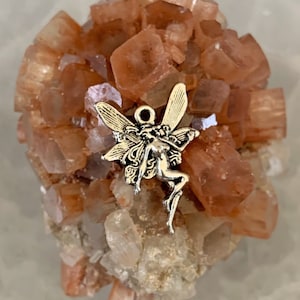 May include: A small, silver fairy charm is attached to a cluster of orange and white crystals. The crystals are arranged in a cluster, resembling a small, natural rock formation.