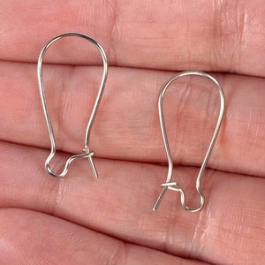 May include: Two silver-colored earring hooks. Each hook has a curved top and a looped bottom with a small post. The hooks are designed to be inserted into pierced ears and secure earrings.
