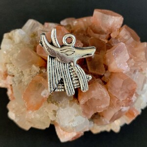 May include: Silver charm of an Egyptian Anubis jackal head, with detailed features, resting on a cluster of pink and white crystals.