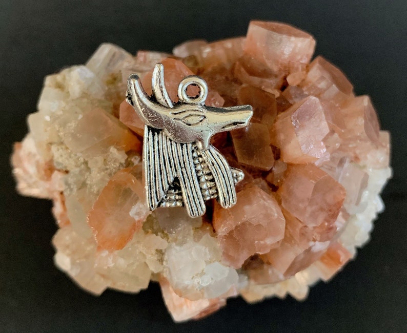 May include: Silver charm of an Egyptian Anubis jackal head, with detailed features, resting on a cluster of pink and white crystals.