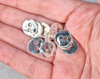May include: A collection of silver-toned metal buttons with two holes each. The buttons are round and have a slightly raised surface.